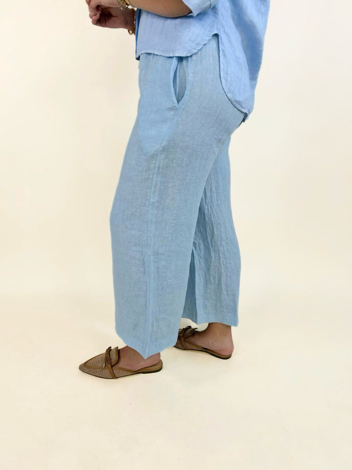 Light blue linen cropped wide-leg pants highlighting the relaxed silhouette, lightweight fabric, side pocket detail, and easy pull-on fit paired with woven pointed-toe mule flats.