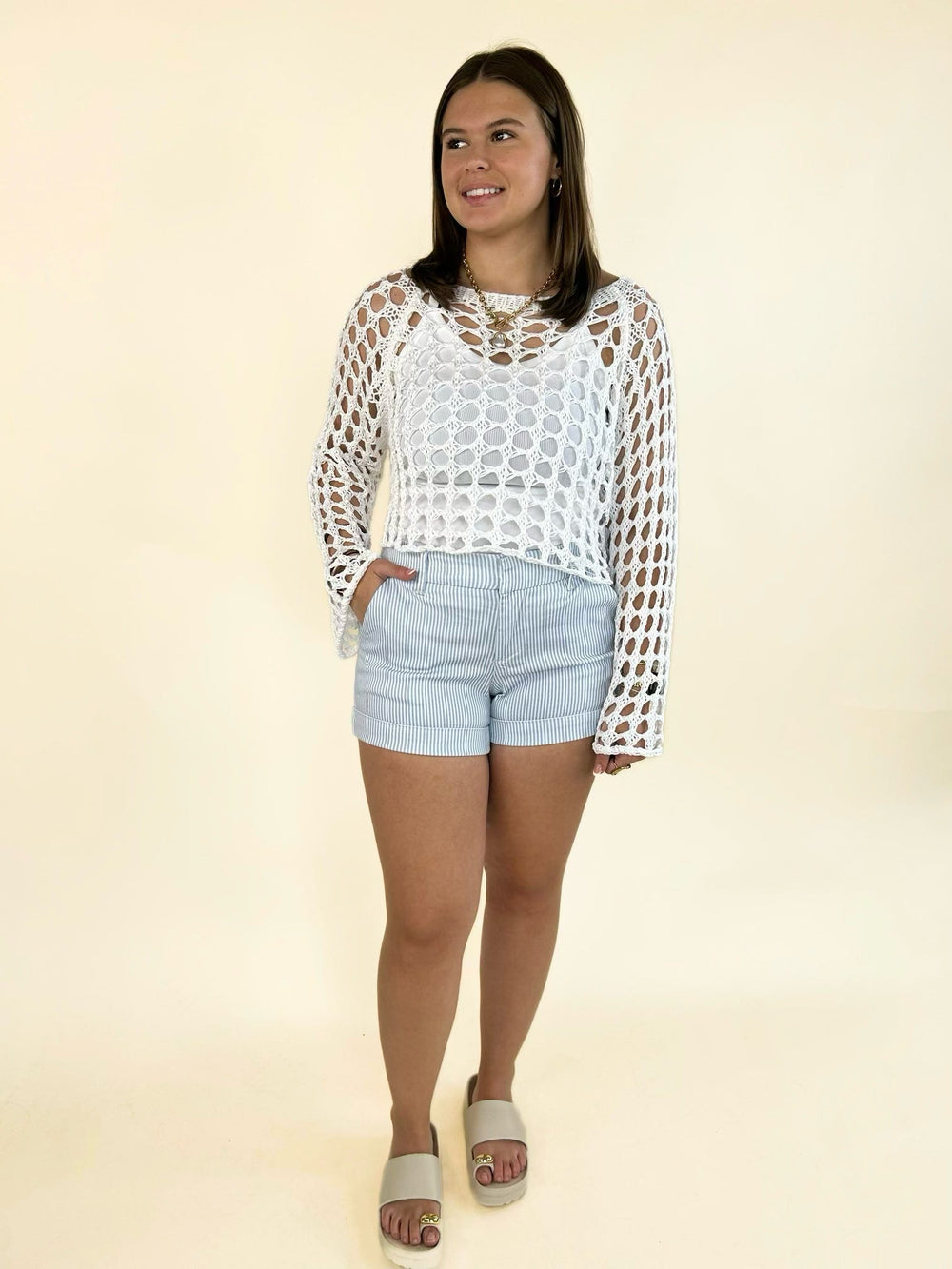 Women's light blue and white pinstripe cuffed shorts with a tailored fit, front pockets, belt loops, and a zip-front closure, styled with a white open-knit crochet sweater and slide sandals.