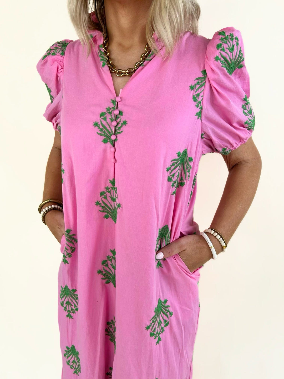 Close-up front view of the pink embroidered midi dress highlighting the V-neckline, covered button-front detail, puff sleeves, green floral embroidery, and side pockets.