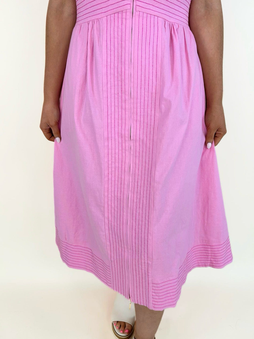 Close-up of the lower half of the pink midi dress featuring pintuck detailing down the center, a flowy skirt, and decorative stitched hem paired with white heeled slide sandals.