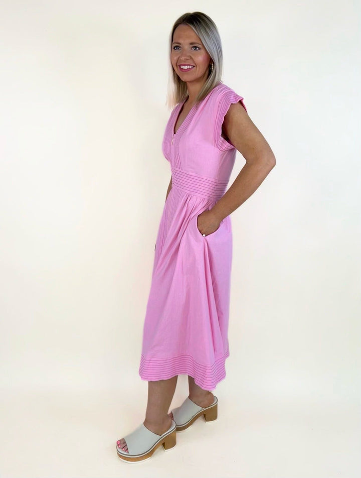 Side view of a pink sleeveless midi dress featuring a V-neckline, front zipper detail, pintuck accents, fitted waist, side pockets, and a flowy skirt styled with white heeled slide sandals.