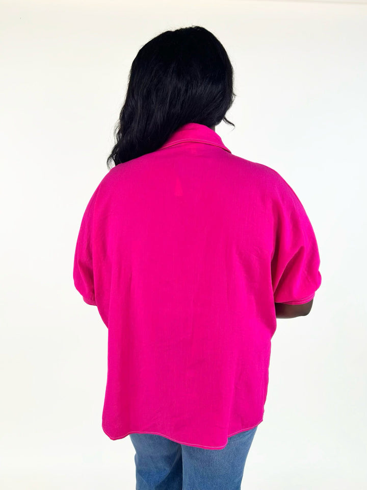 Back view of the bright fuchsia relaxed fit blouse with short cuffed sleeves, collared neckline, and slightly curved hem.