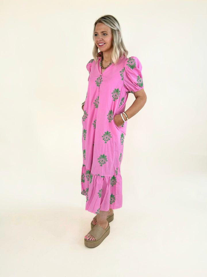 Side view of model wearing a pink embroidered midi dress with green floral embroidery, puff sleeves, button-front detail, side pockets, and a tiered ruffle hem paired with platform slide sandals.