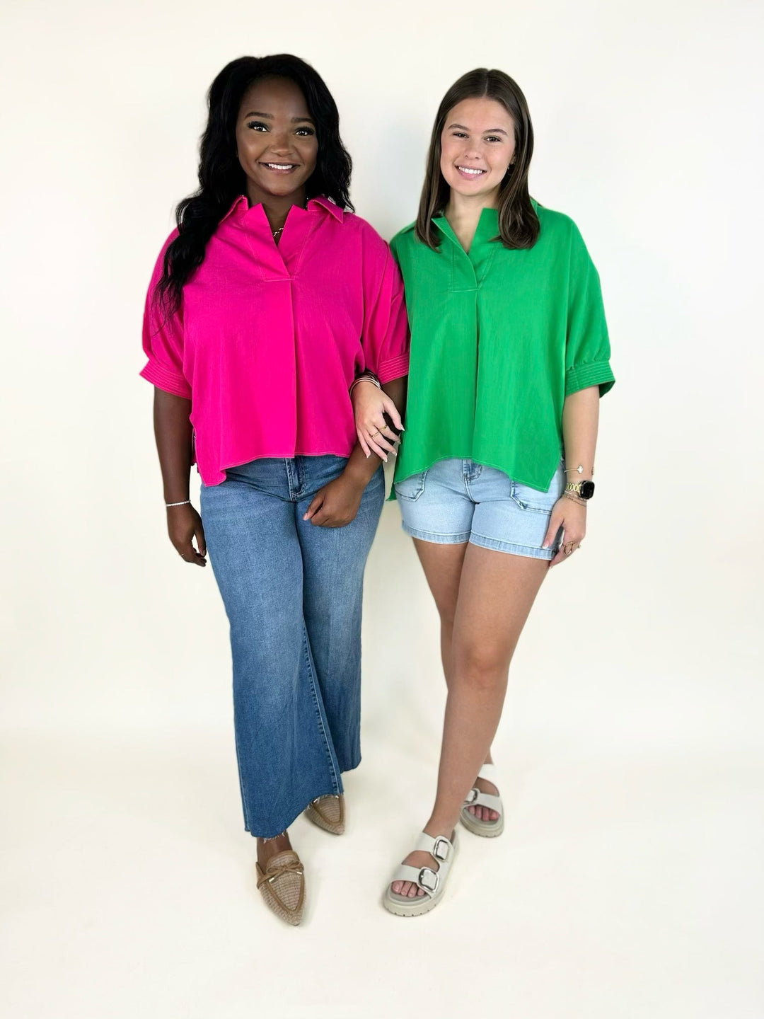 Model wearing a bright fuchsia collared short sleeve blouse with relaxed fit and light wash wide leg jeans standing beside a model wearing the same blouse in vibrant green with light wash denim shorts.