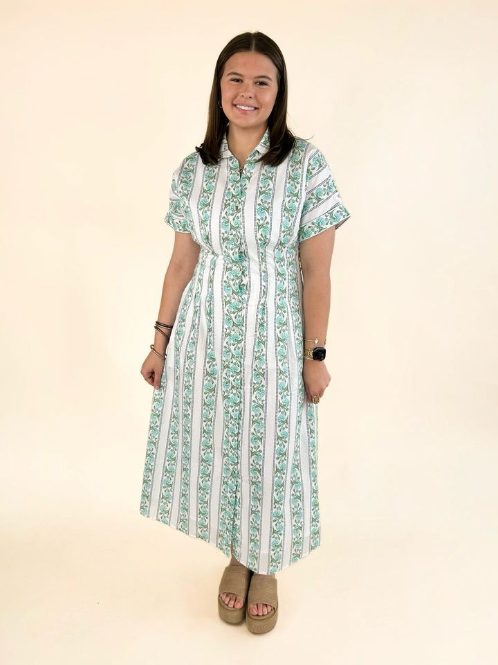 Front view of model wearing a white collared button-front midi dress with green floral stripe print, short sleeves, fitted waist, side pockets, and tan platform sandals.