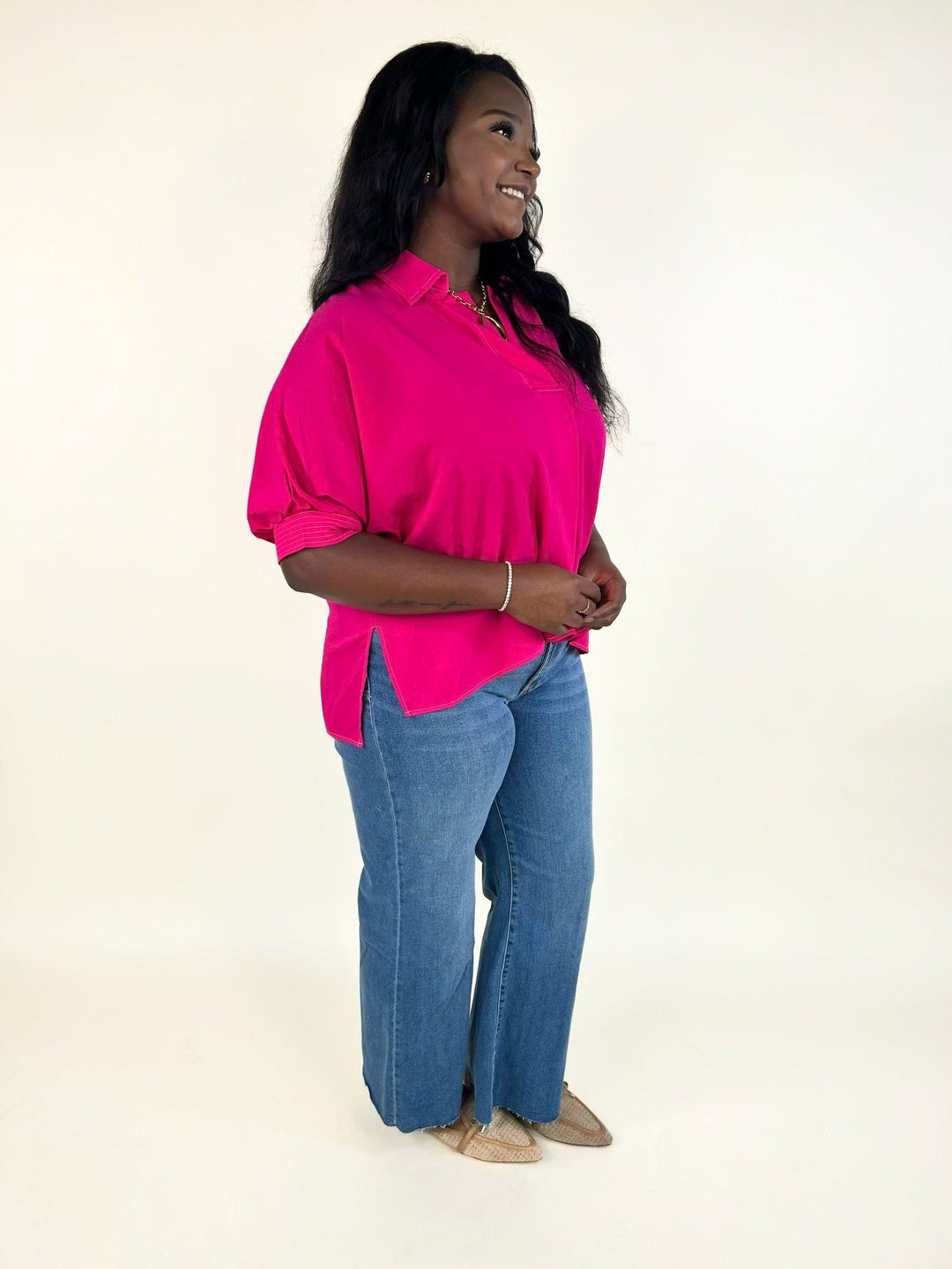 Side view of the bright fuchsia collared blouse featuring a relaxed silhouette, side slit hem, lightweight woven fabric, and cuffed short sleeves styled with wide leg jeans.