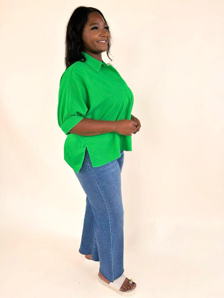 Side profile of the vibrant green collared blouse with relaxed fit, side slit hem, cuffed short sleeves, and lightweight woven fabric paired with light wash jeans.