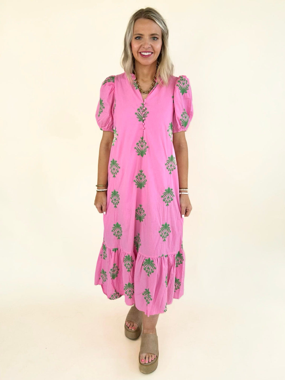 Front view of model wearing a pink midi dress with green floral embroidery, puff sleeves, covered button-front detail, relaxed fit, side pockets, and a tiered ruffle hem.