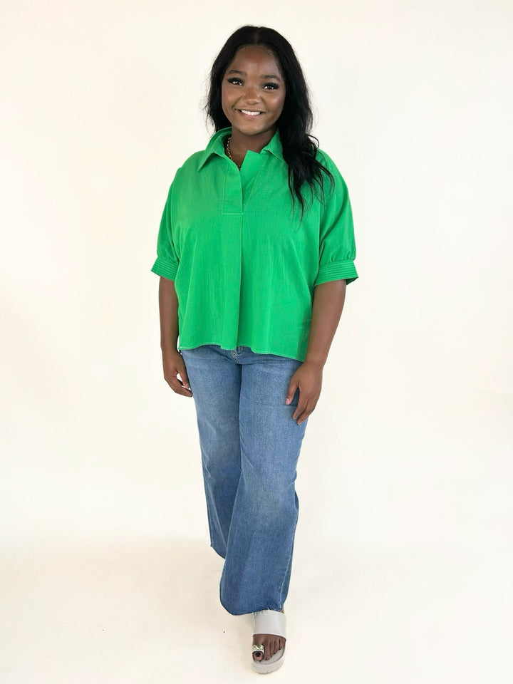 Front view of model wearing the vibrant green collared short sleeve blouse with a relaxed fit, split neckline, side slits, and light wash wide leg jeans.