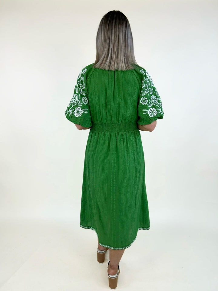 Back view of a green embroidered midi dress with white floral embroidered puff sleeves, textured woven fabric, a smocked waist, and a flowy midi-length silhouette.