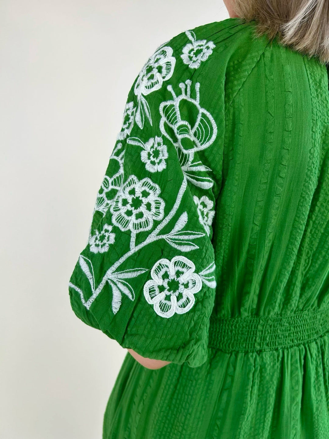 Close-up of the back shoulder of a green midi dress featuring white floral embroidery on the puff sleeve, textured woven fabric, and a smocked waist detail.