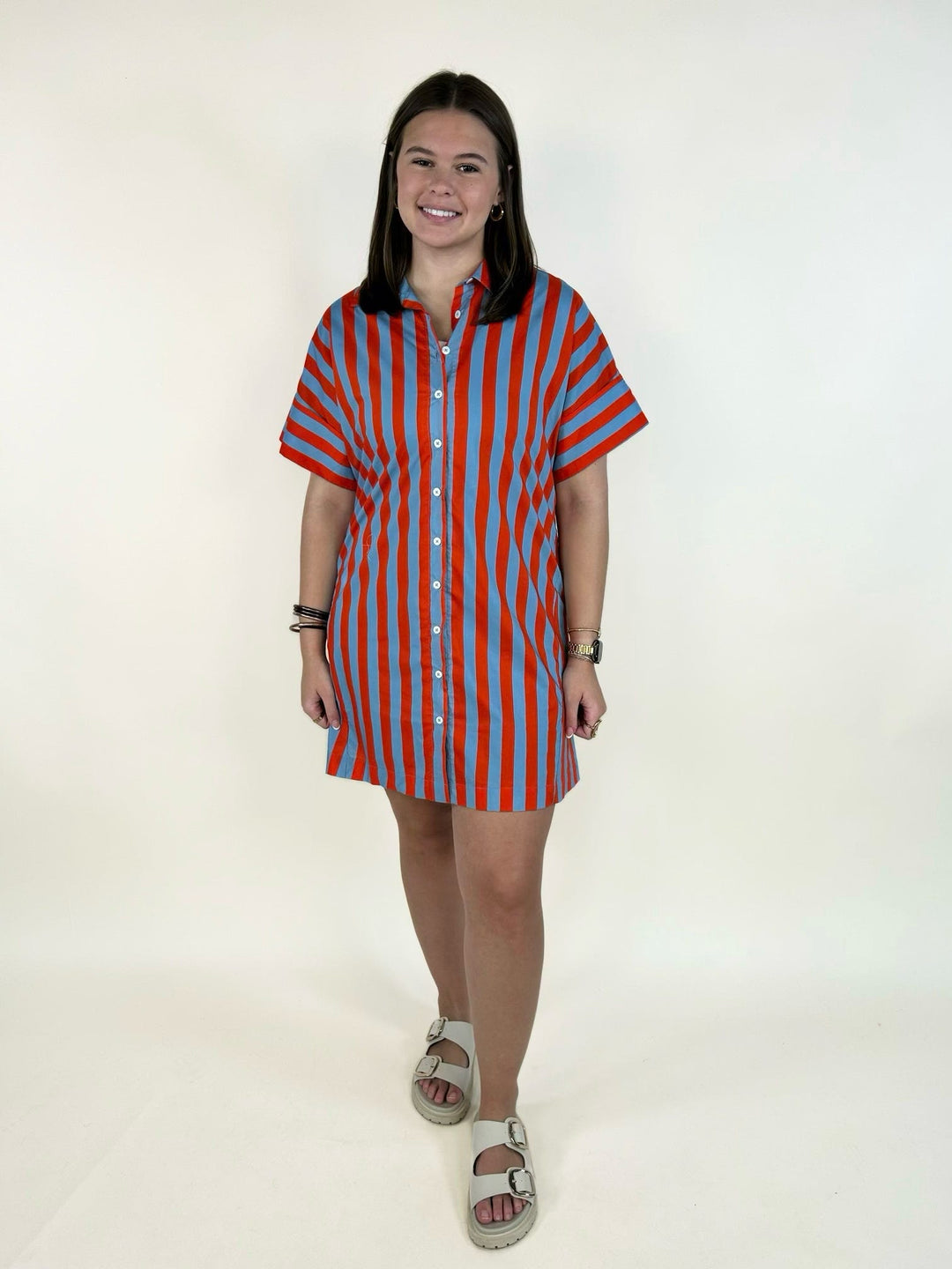 Model wearing the THML Weekend Getaway Striped Shirt Dress in blue with orange vertical stripes, featuring a collared neckline, button-front closure, rolled short sleeves, side pockets, and a relaxed mini-length silhouette.