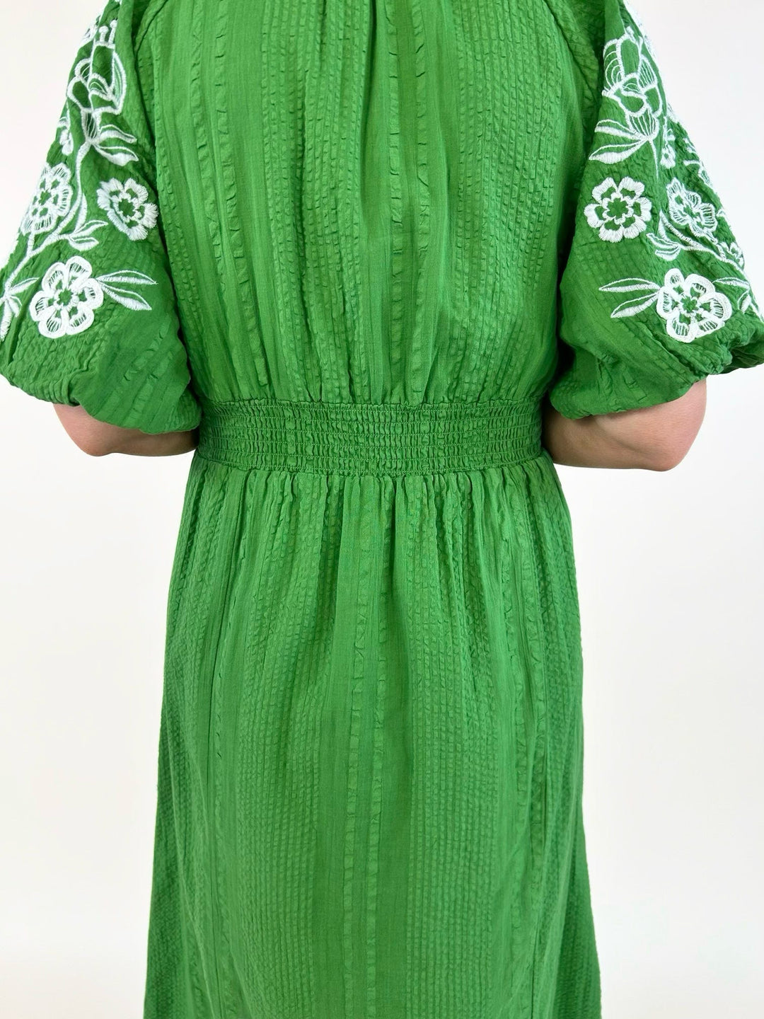 Back view close-up of a green textured midi dress showcasing the smocked elastic waist, white floral embroidered puff sleeves, and lightweight woven fabric.