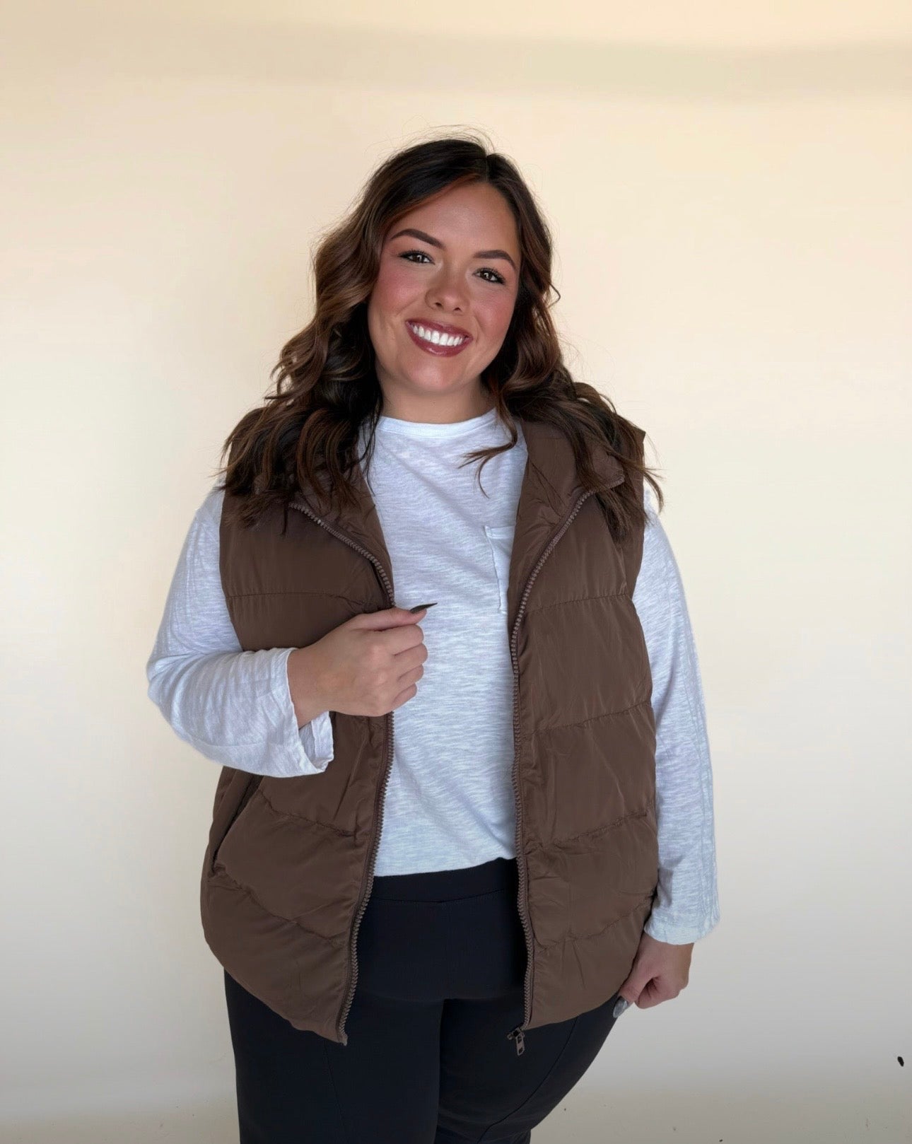 Woman wearing a chocolate brown puffer vest over a white long-sleeve top, smiling with one hand near the vest opening against a simple neutral backdrop.