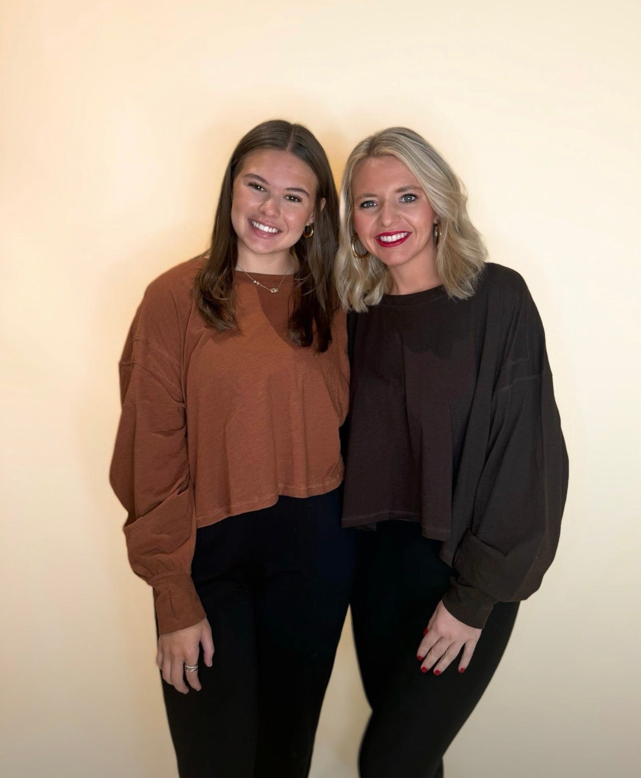 Two women wearing oversized long sleeve tops, one in warm brown and one in dark chocolate brown, both paired with black pants for a casual fall outfit.