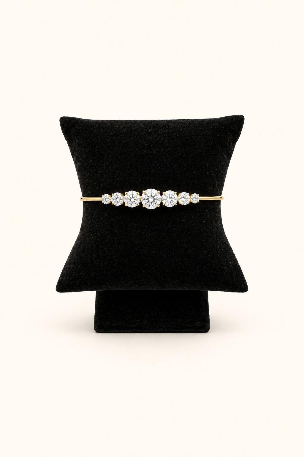 A gold bracelet with a row of clear gemstones, displayed on a black cushion stand against a light cream background.