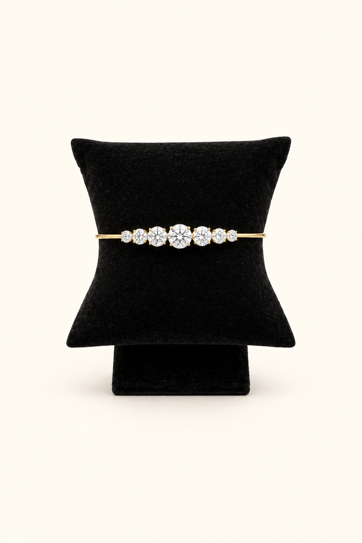 A gold bracelet with a row of clear gemstones, displayed on a black cushion stand against a light cream background.