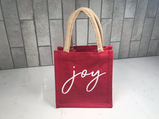 Holiday mini tote bag in red woven fabric featuring the word joy in white script, standing upright against tile background.
