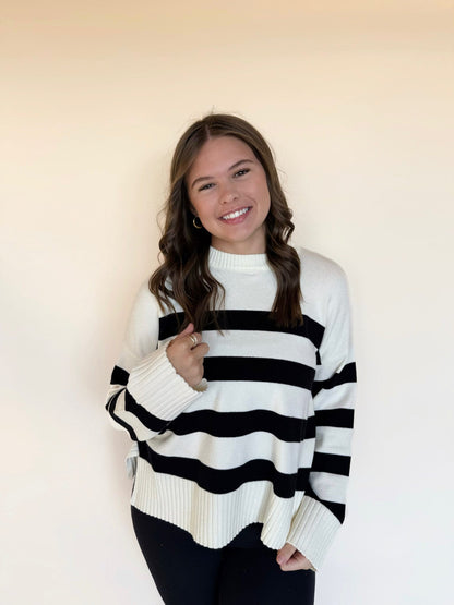 Woman wearing an oversized ivory and black striped knit sweater with a mock neck and long ribbed sleeves, styled with black leggings against a neutral background.