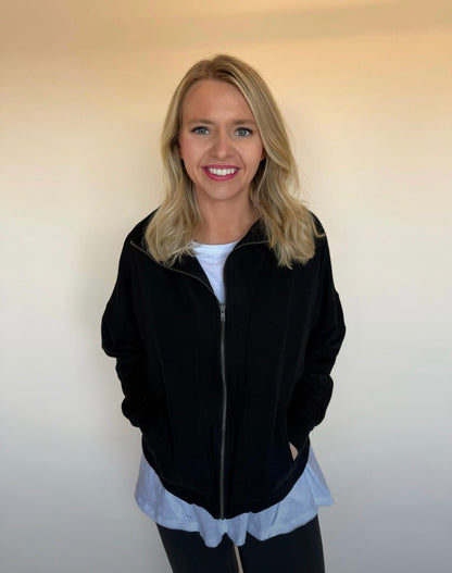 Woman wearing a black jacket over a white shirt with a white shirt underneath, standing against a plain background.