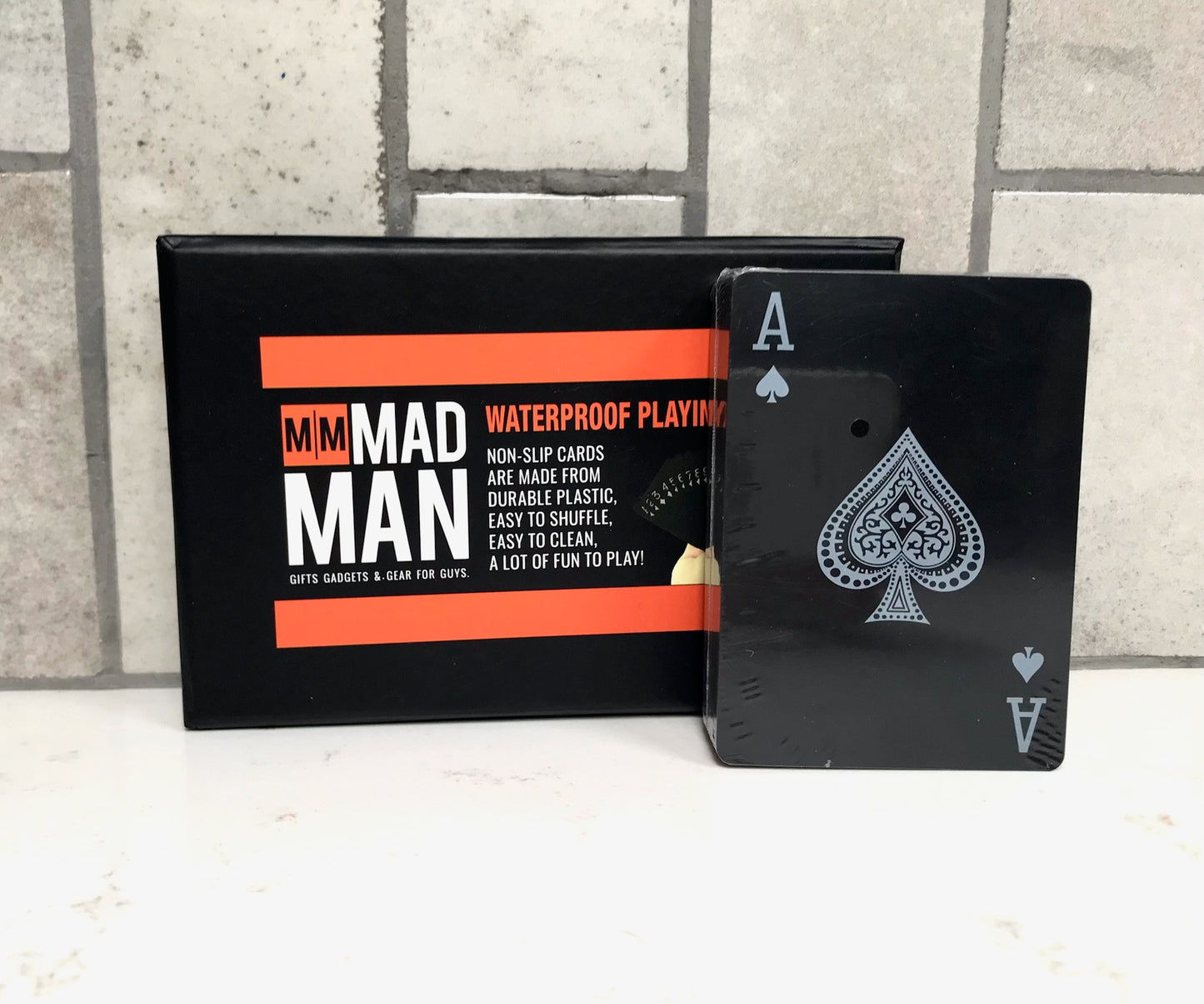 Black playing cards with Ace of Spades design and packaging labeled 'Mad Man' on a tiled floor background.