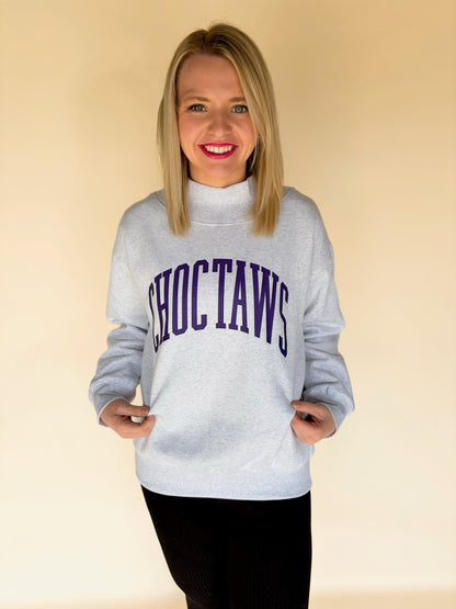 Woman wearing light gray Choctaws mock neck sweatshirt with purple varsity lettering, cozy women’s boutique college style pullover