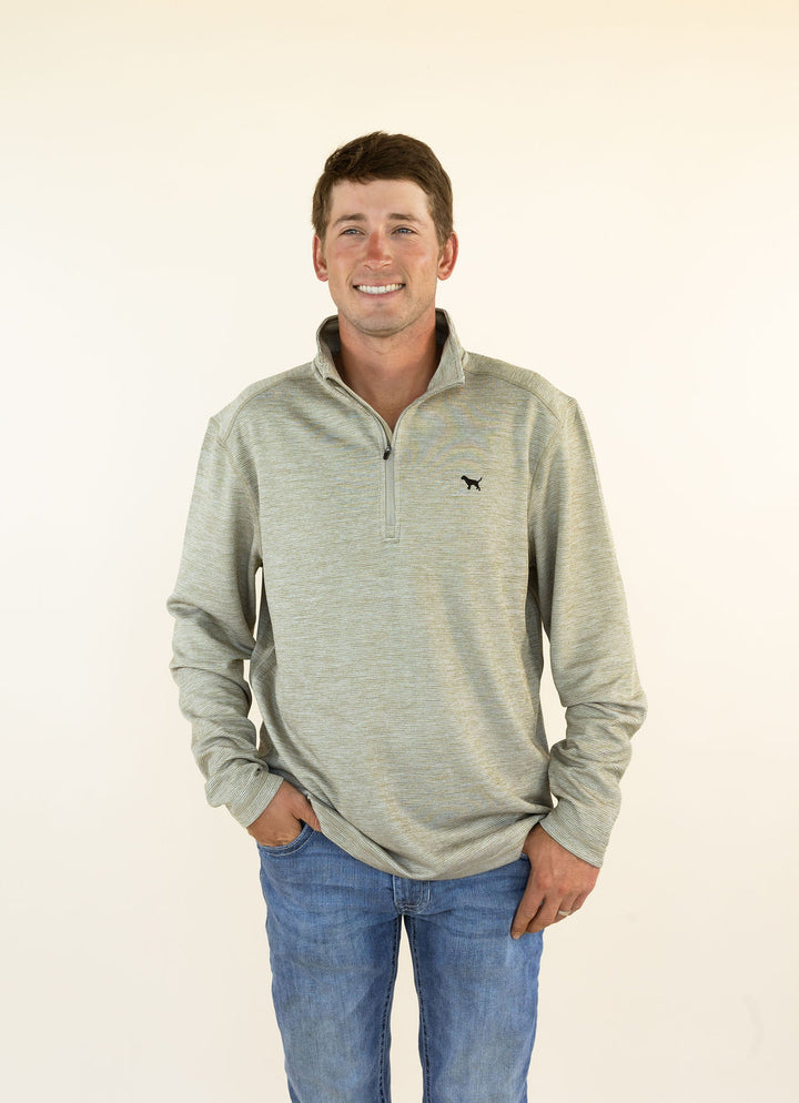 A smiling man wearing a light heather olive quarter-zip pullover sweater with a small black dog logo on the left chest, paired with blue jeans, standing against a plain off-white background.