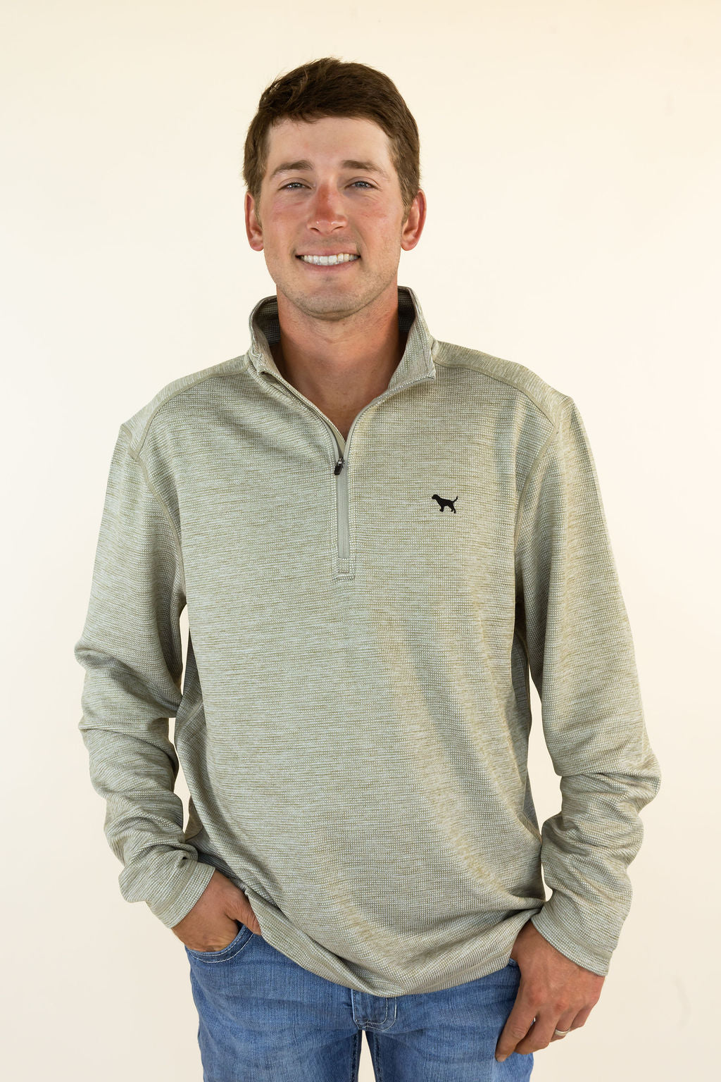 A man wearing a light heather olive quarter-zip pullover with a small black dog logo on the left chest, paired with blue jeans, standing against a plain off-white background.