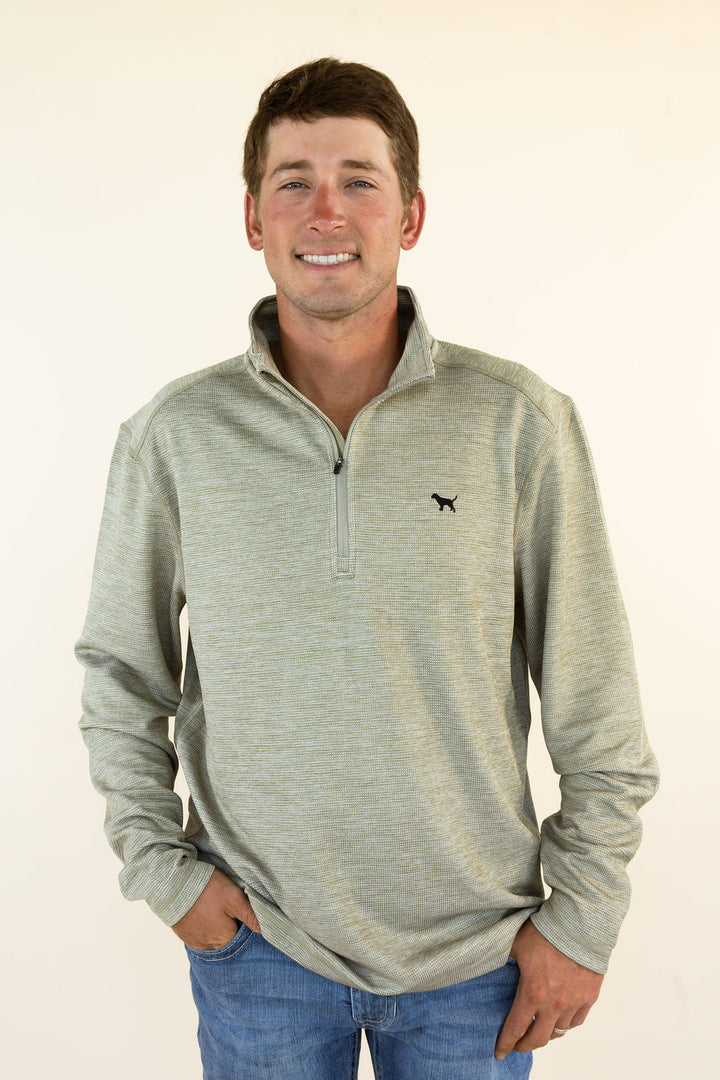 A man wearing a light heather olive quarter-zip pullover with a small black dog logo on the left chest, paired with blue jeans, standing against a plain off-white background.