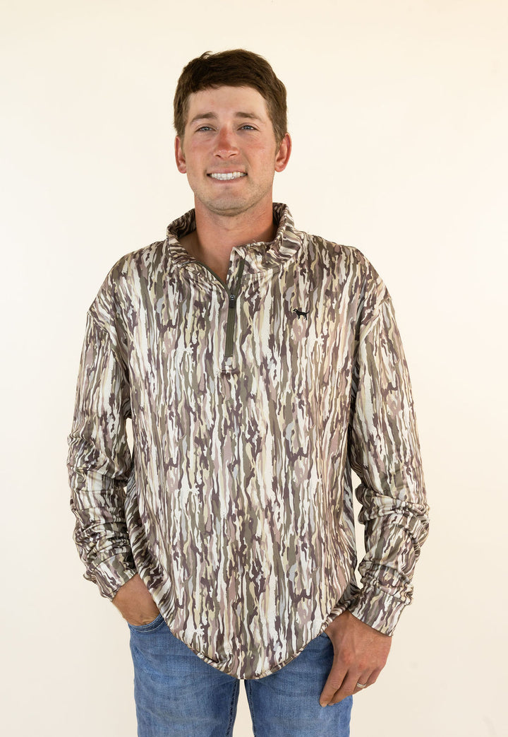 A man wearing a long-sleeve button-down shirt with a vertical bark camouflage pattern in brown, beige, and gray, paired with blue jeans, standing against a plain off-white background.