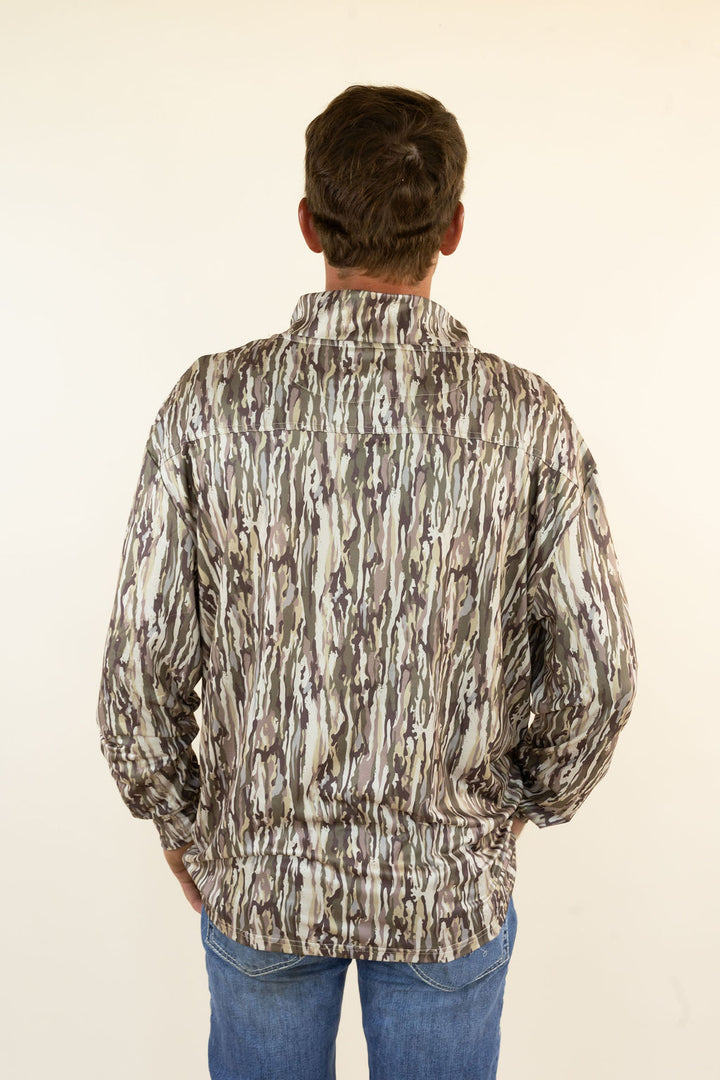 A man viewed from behind wearing a long-sleeve button-up shirt with a brown, gray, and beige vertical bark camouflage pattern, paired with blue jeans, against a plain off-white background.