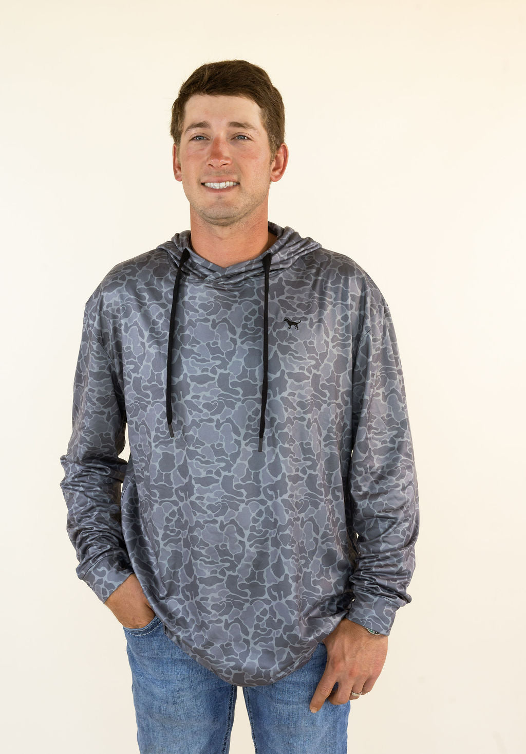 A man wearing a gray camouflage hooded long-sleeve shirt with blue jeans, standing against a plain off-white background.
