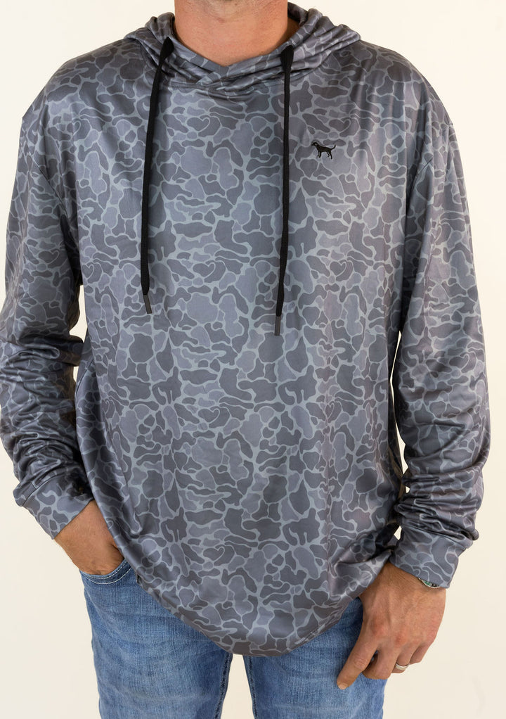 A man wearing a gray camouflage-patterned hooded long-sleeve shirt with black drawstrings, paired with blue jeans, against a plain light background.