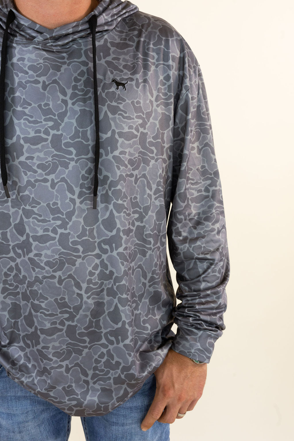 A close-up of a person wearing a gray camouflage patterned hooded long-sleeve shirt with black drawstrings and a small black dog logo on the left chest, paired with blue jeans against a plain light background.