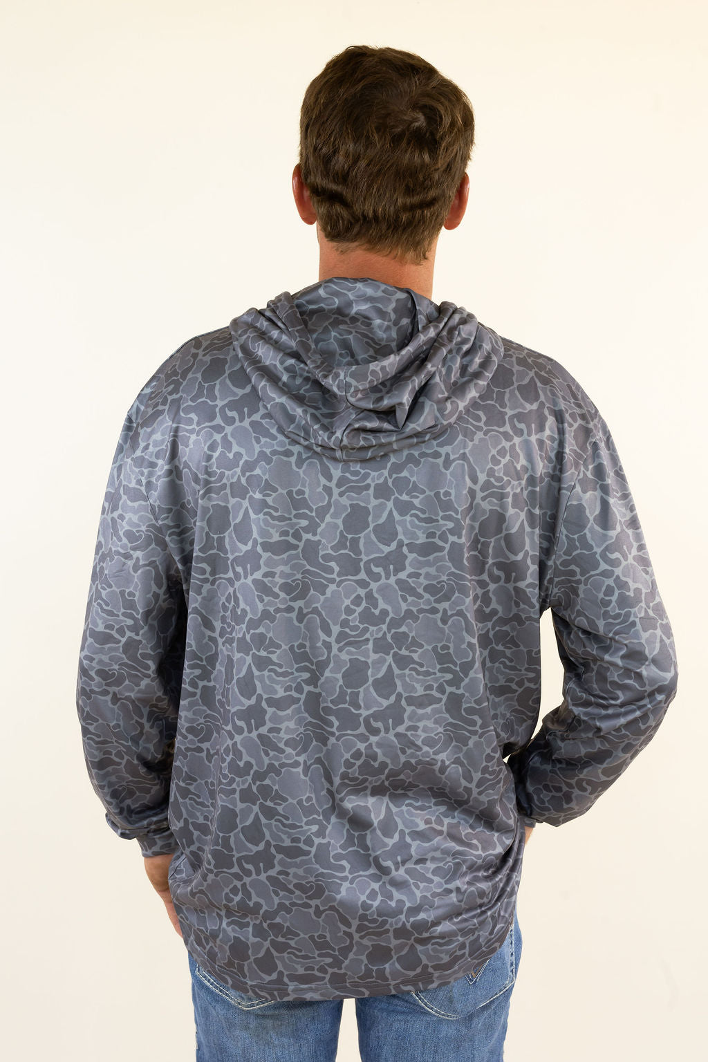 A man viewed from behind wearing a gray hooded sweatshirt with a dark gray camouflage pattern, paired with blue jeans, against a plain off-white background.