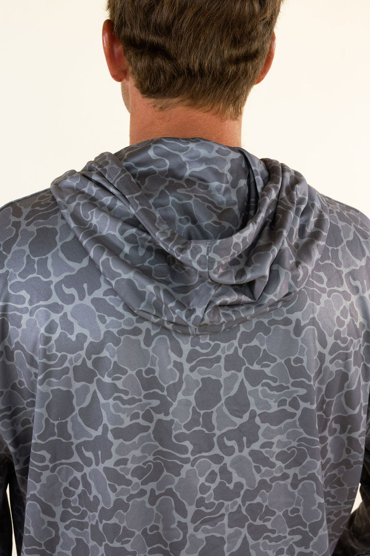 Back view of a person wearing a gray hooded shirt with a dark gray camouflage pattern, against a plain off-white background.