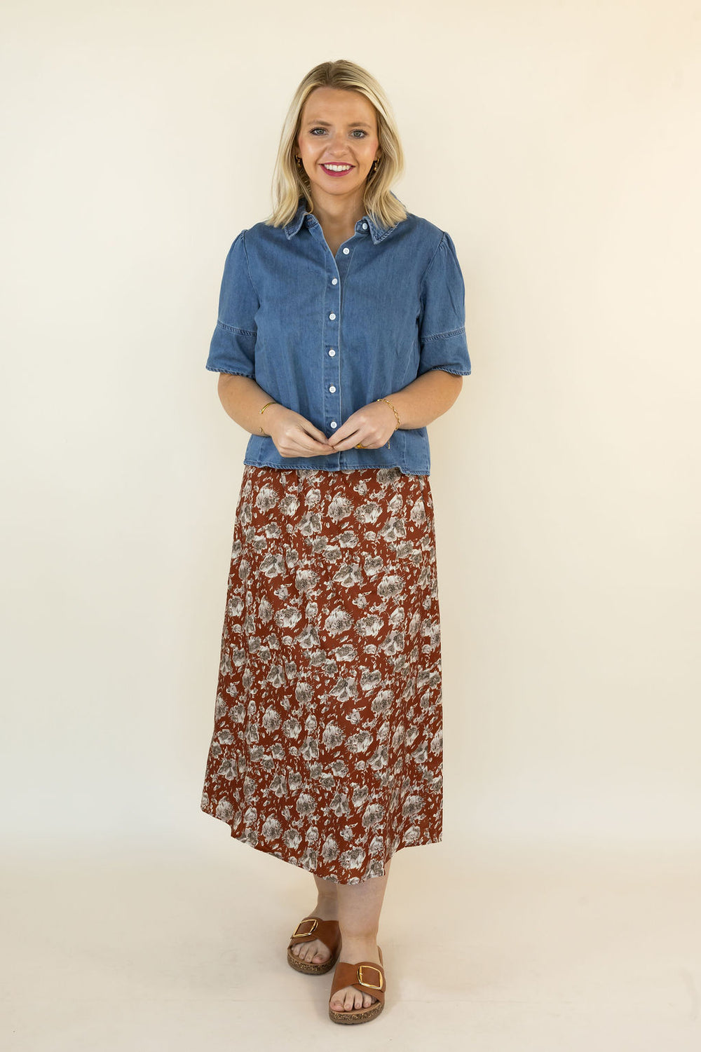 A woman modeling a two-piece outfit consisting of a blue short-sleeve button-up shirt and a rust-brown midi skirt with a white floral pattern, paired with brown sandals, against a plain cream background.