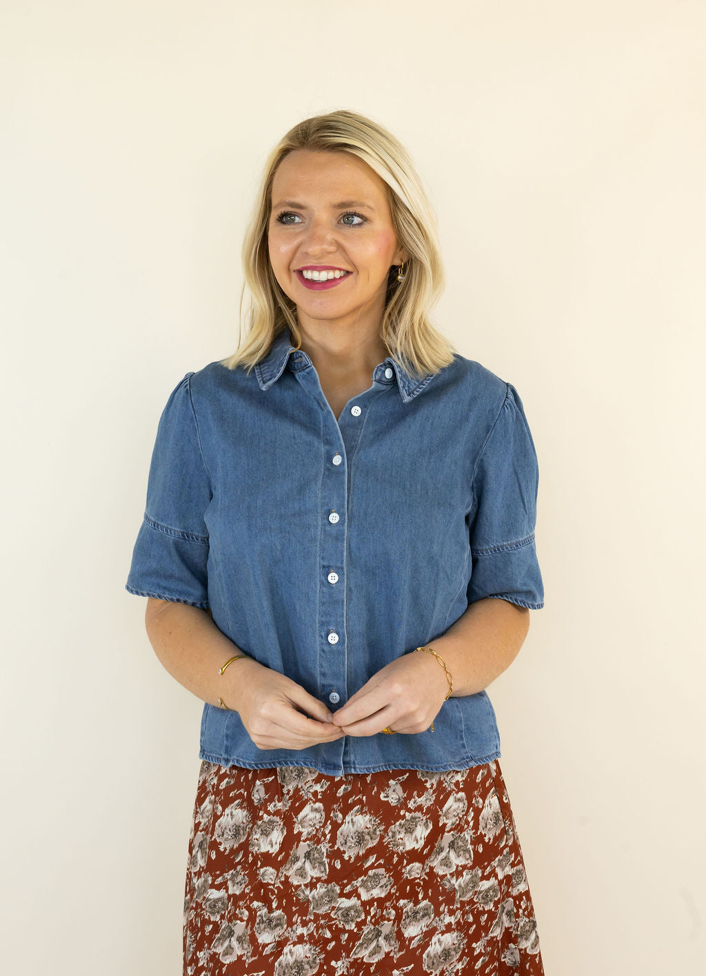 A smiling blonde woman wearing a short-sleeve blue denim button-up shirt and a rust-colored floral skirt, standing against a plain off-white background.
