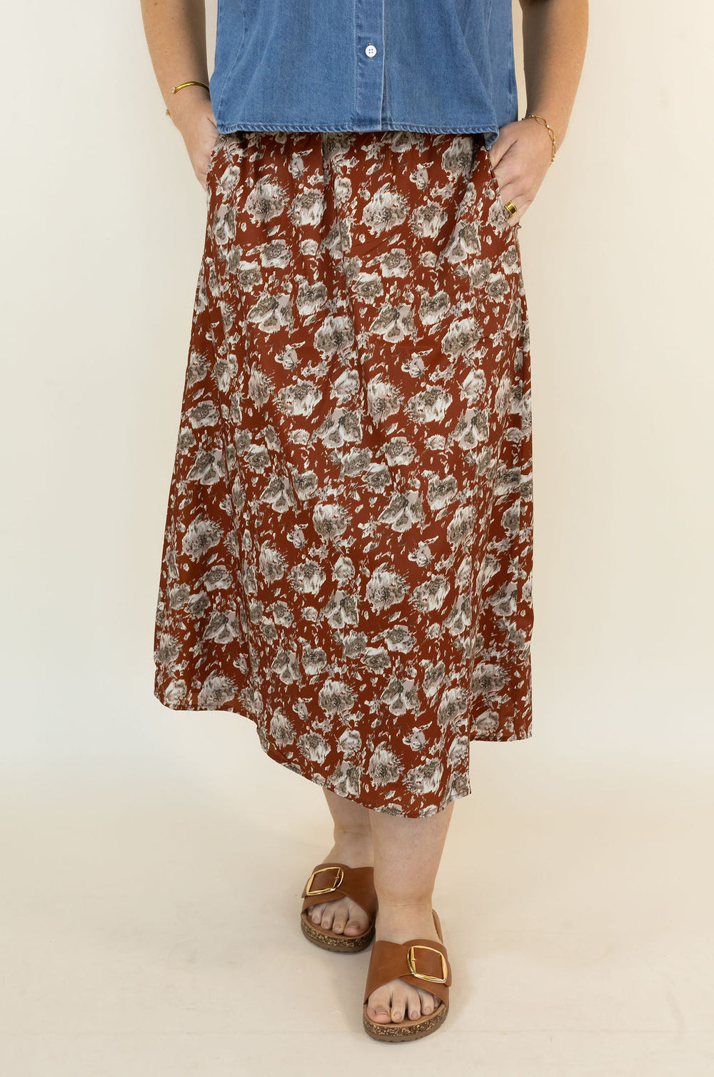 A person wearing a hickory midi skirt with a gray and white floral pattern, paired with a blue denim shirt and brown slide sandals, standing against a plain off-white background.
