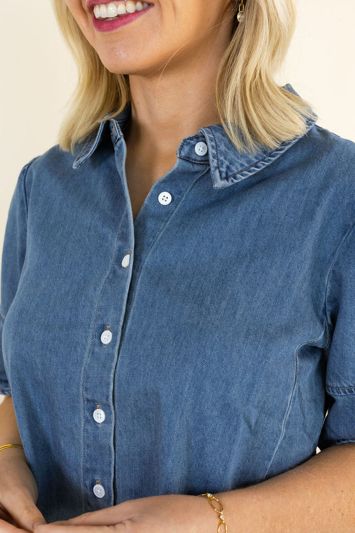 A close-up of a woman wearing a blue denim button-up shirt, showing the collar, front buttons, and part of her blonde hair and hands against a light beige background.