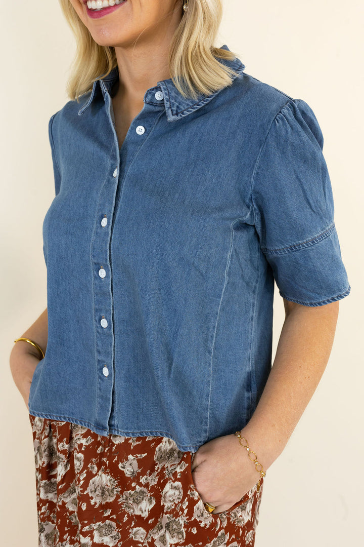 A woman wearing a short-sleeve blue denim button-up shirt with puff sleeves and a brown floral skirt, photographed against a plain light beige background.