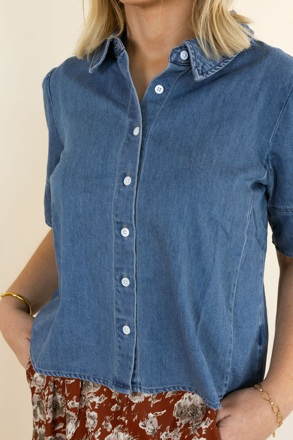 A close-up of a woman wearing a blue denim short-sleeve button-up shirt with a pointed collar and white buttons, paired with a rust-colored floral skirt.