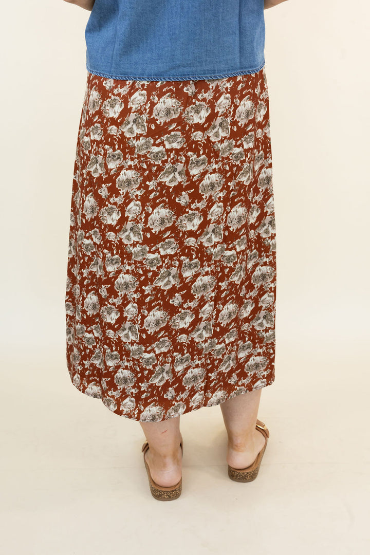 A person shown from the waist down wearing a hickory midi skirt with a gray and white floral pattern, paired with a blue top and brown wedge sandals, against a plain beige background.