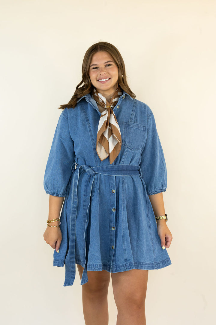 A woman modeling a blue denim shirt dress with a tie-neck and button-front detail, styled with a brown and white patterned neck scarf, against a plain cream background.