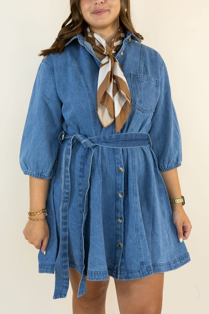 A woman wearing a blue denim shirt dress with puff sleeves, a tie waist belt, and front buttons, styled with a brown and white patterned neck scarf, against a plain cream background.