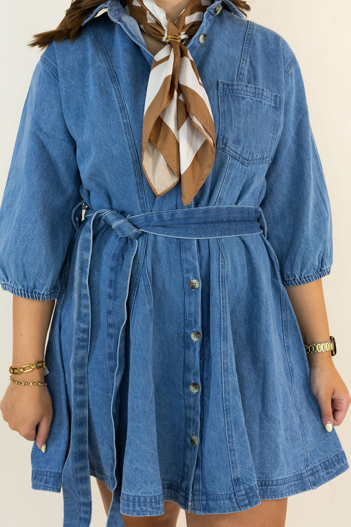 A person wearing a blue denim shirt dress with a tie waist belt and front button closure, styled with a brown and cream patterned neck scarf. The photo is cropped from the shoulders to the upper thighs against a plain light beige background.