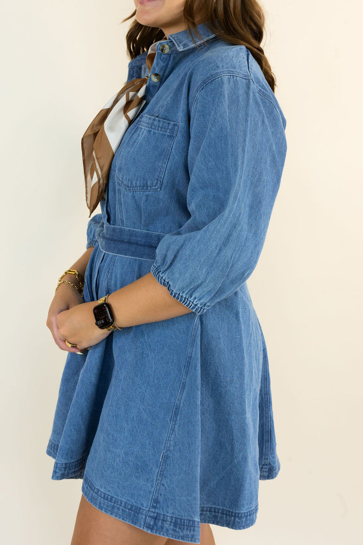 A woman wearing a blue denim shirt dress with three-quarter sleeves, a buttoned collar, and a matching waist tie, styled with a brown patterned scarf against a plain cream background.