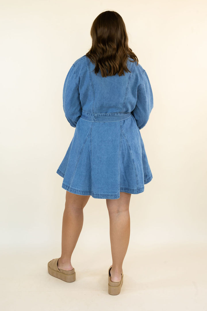 A woman viewed from behind wearing a light blue denim dress with long puff sleeves and a flared skirt, paired with tan platform clogs, against a plain cream background.