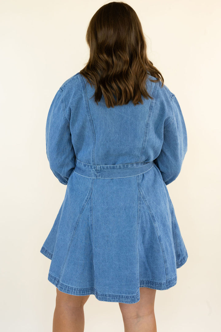 A woman viewed from behind wearing a light blue denim belted dress with long puff sleeves, against a plain cream background.