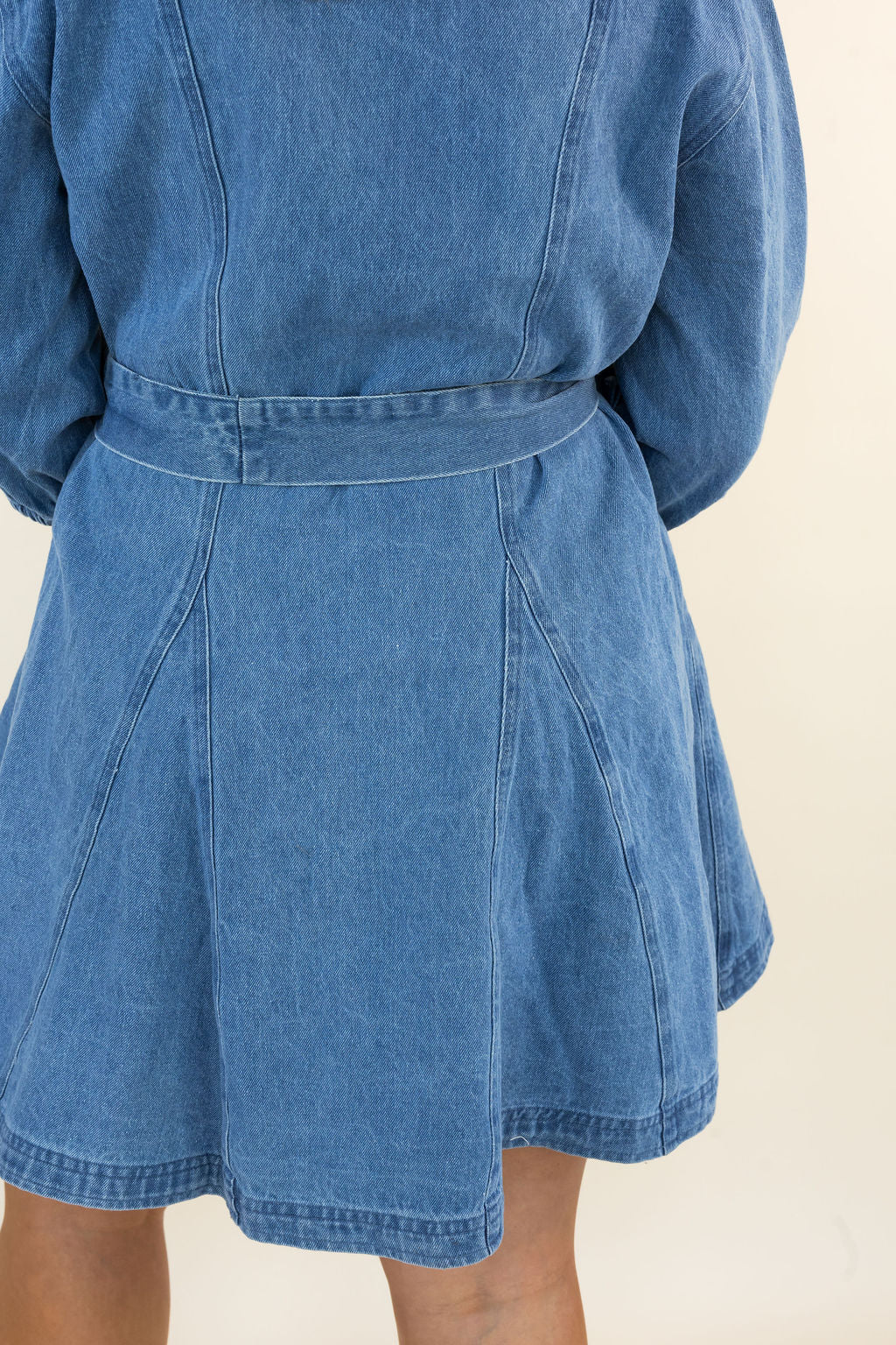 Close-up back view of a person wearing a blue denim belted dress with a flared skirt, against a plain light beige background.